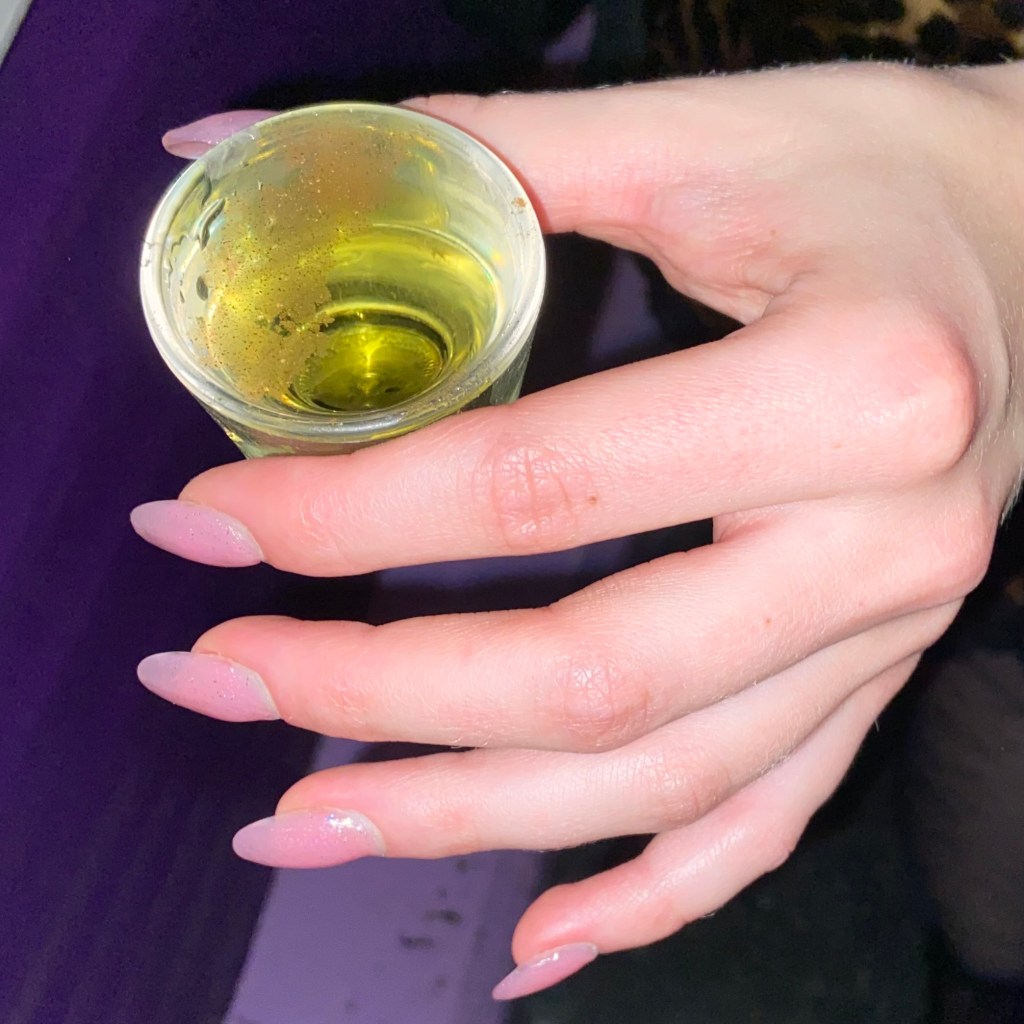 Hand with pink medium length oval nails holding a shot glass with green liquid.
