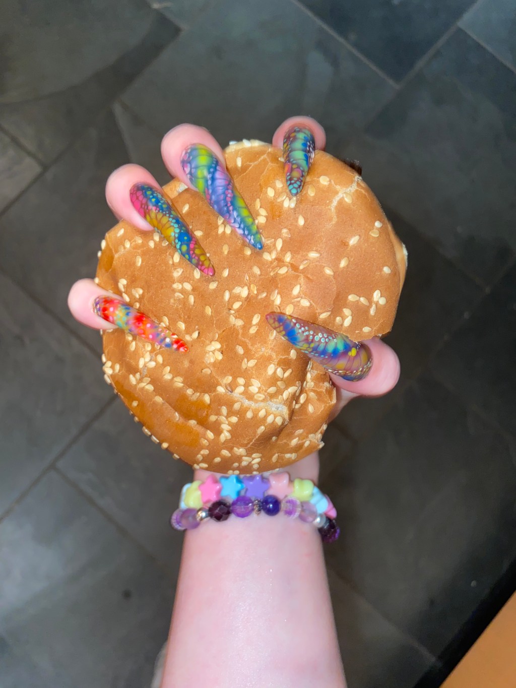Hand with xxl stiletto shaped multicolour blooming gel nails gripping a burger.