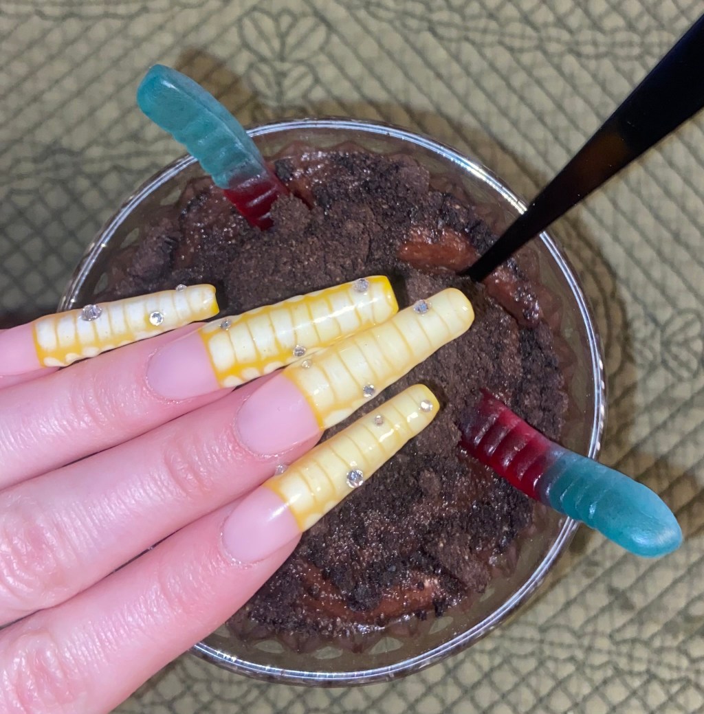 Yellow French Tip nails with the crocodile blooming gel pattern hovering over a cup of dirt and worms pudding.